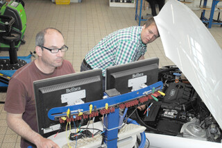 eine Person vor einem PC während eine weitere Person an einem Auto arbeitet eine Person vor einem PC während eine weitere Person an einem Auto arbeitet